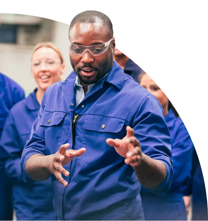 Man in blue overalls and safety glasses explaining with hand gestures to a group