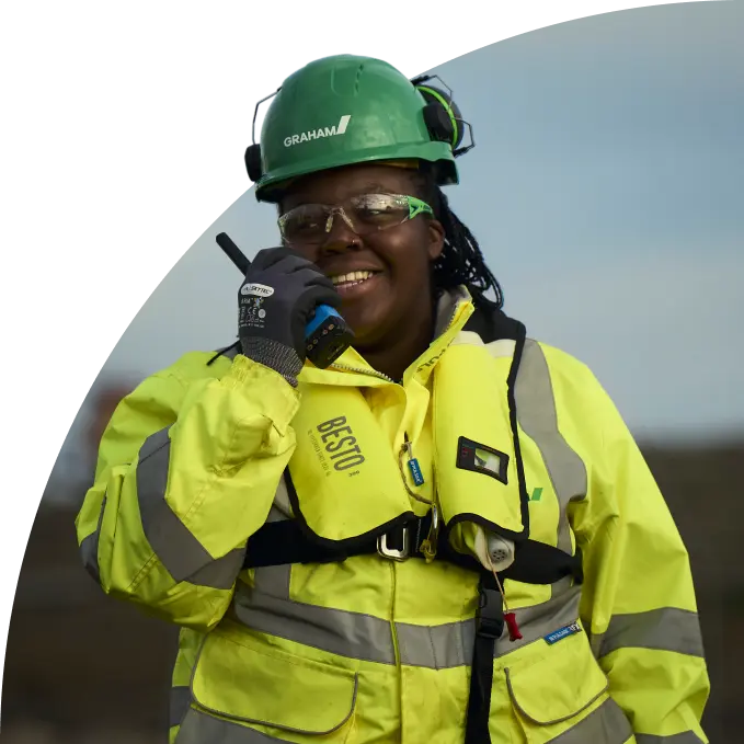 Woman in high visibility safety gear and green hard hat speaking into a walkie-talkie